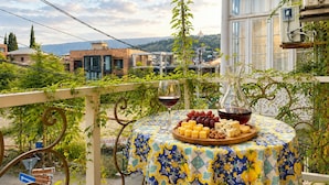 Un balcón con una mesa preparada para una comida, una copa de vino y vistas a edificios y colinas.