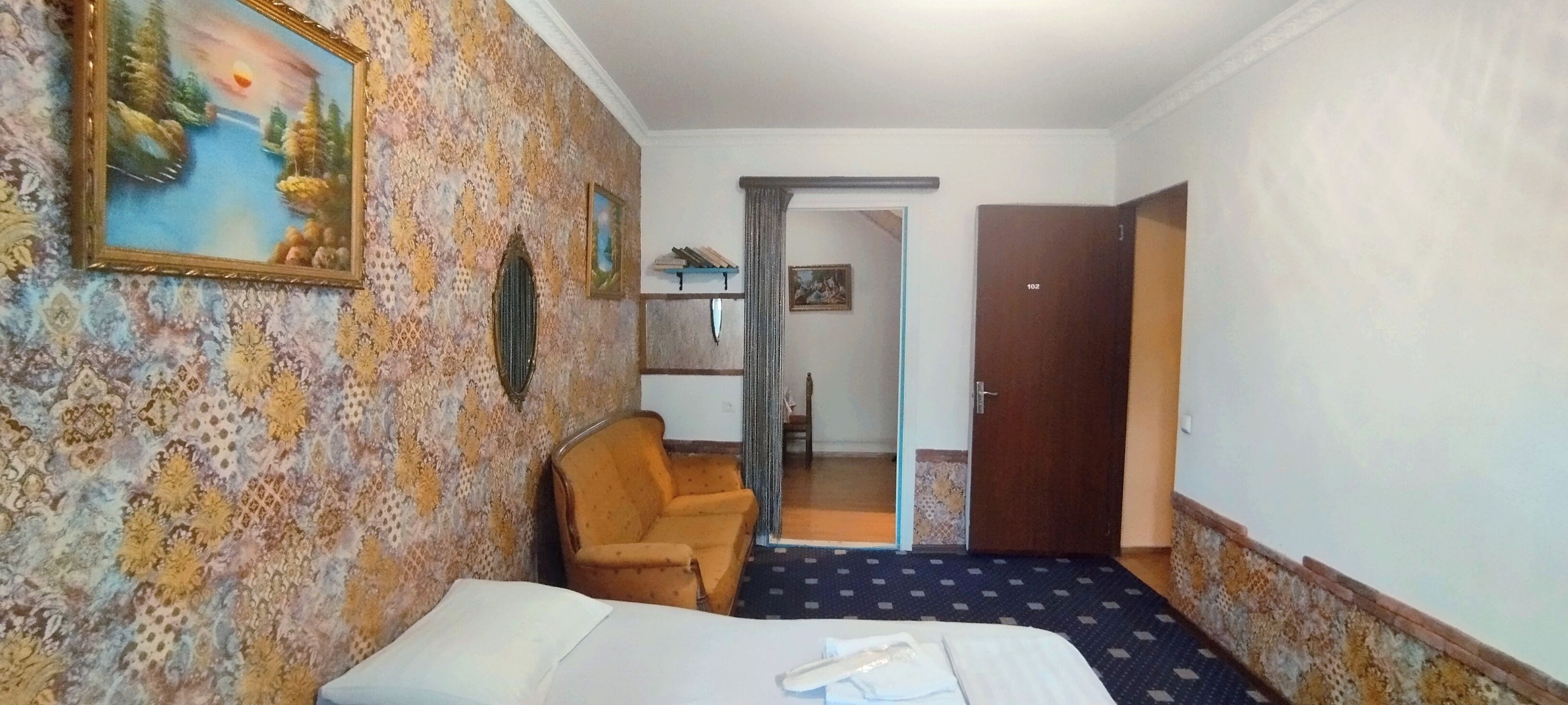 A hotel room with a patterned wallpaper, a bed, a chair, and a door leading to another room.