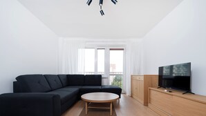 A modern living room with a sectional sofa, a round coffee table, a TV on a wooden stand, and a view of the outdoors through large windows.