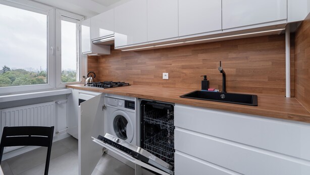 A modern kitchen with white cabinets, a wooden countertop, a built-in washing machine, and a window overlooking a cityscape.