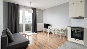 Un salon moderne avec un canapé gris, une table à manger avec des chaises blanches, un téléviseur et un coin cuisine doté de meubles blancs et d’une cuisinière.