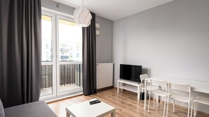 Un salon moderne avec un canapé gris, une table basse blanche et un téléviseur posé sur un meuble. On y trouve des chaises blanches et une petite table avec une télécommande.