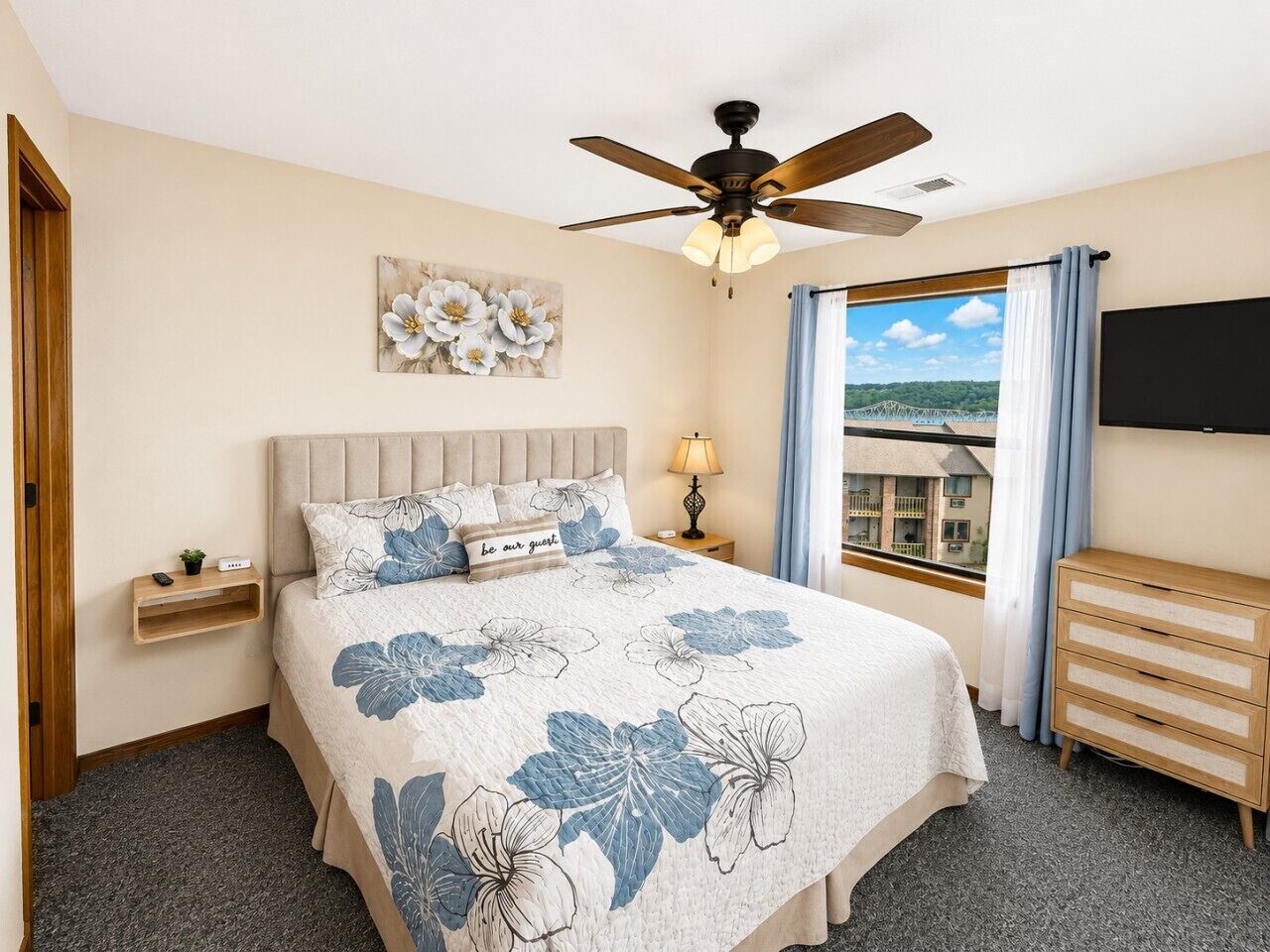 A bedroom with a bed, a ceiling fan, a nightstand, a dresser, a window with a view, and a wall-mounted TV.