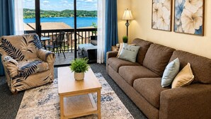 A living room with a sofa, armchair, and a small table with a plant. There is a balcony with a view of a lake and hills. - Pleasure Point at Kimberling Crossing (Kimberling City)