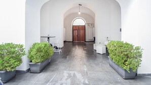 A hallway with a tiled floor, white walls, and a wooden door at the end. There are two potted plants on either side of the hallway.