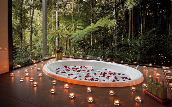 Un bain à remous circulaire, entouré de bougies et de pétales de roses, se dressant sur un fond de verdure luxuriante et d’arbres imposants.