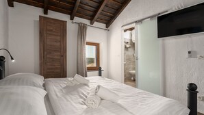 A bedroom with a wooden ceiling, a bed with white linens, a TV mounted on the wall, and a bathroom visible through an open door. - Villa Nina (Istria - Buje) (Buje)