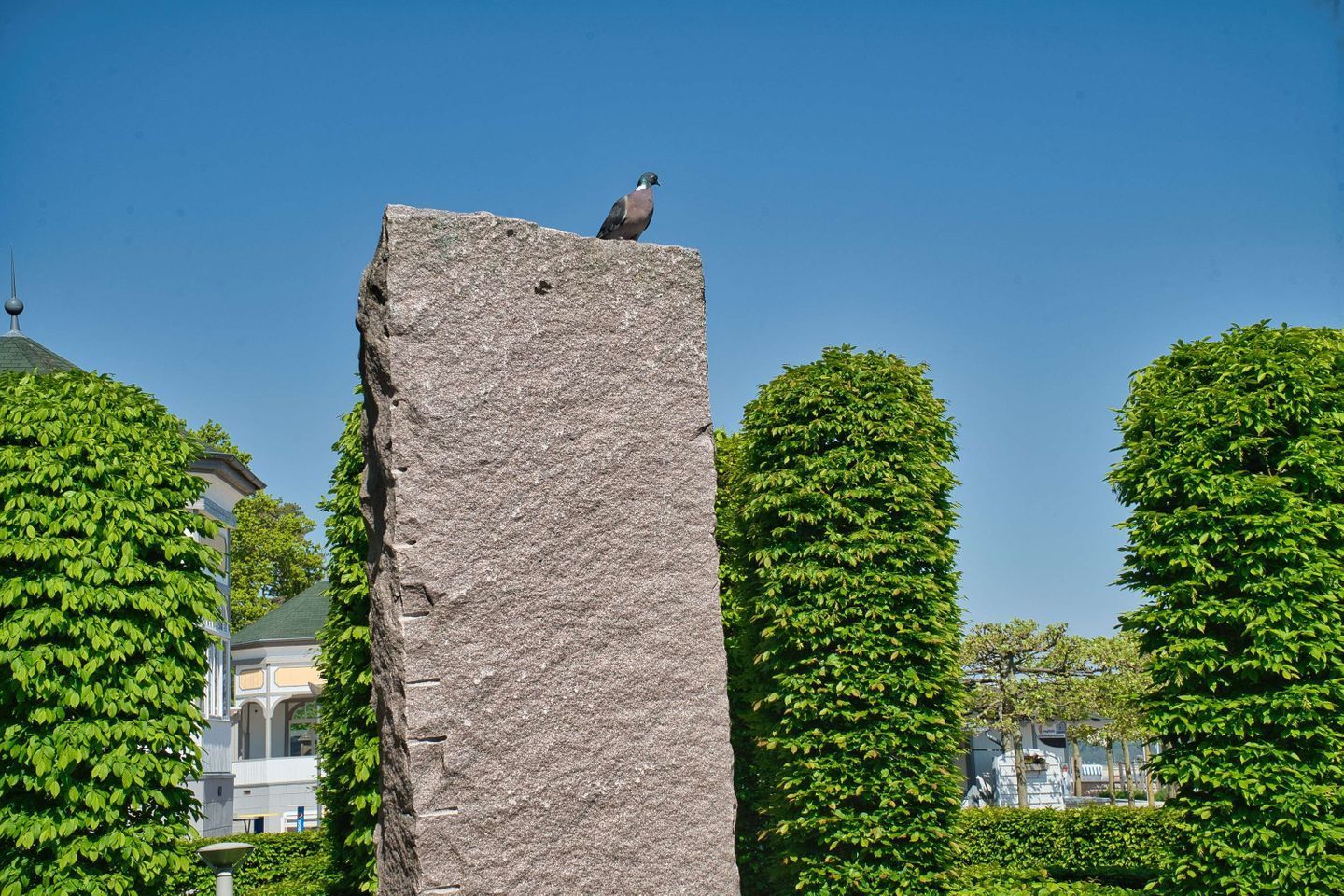 A tall, rectangular stone monument with a bird perched on top, surrounded by well-maintained green hedges and a building with a dome in the background.
