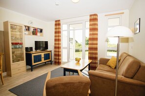 A living room with a sofa, a coffee table, a TV, a cabinet with glass doors, and curtains. - Terrace, free use of the AHOI adventure pool and sauna - Sea view apartment 02 - (Göhren)