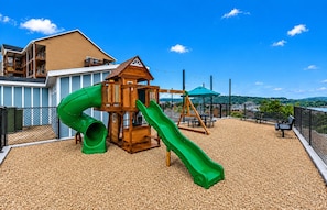 Une aire de jeux avec un toboggan et une structure d’escalade, entourée d’une clôture et offrant une vue sur une étendue d’eau.