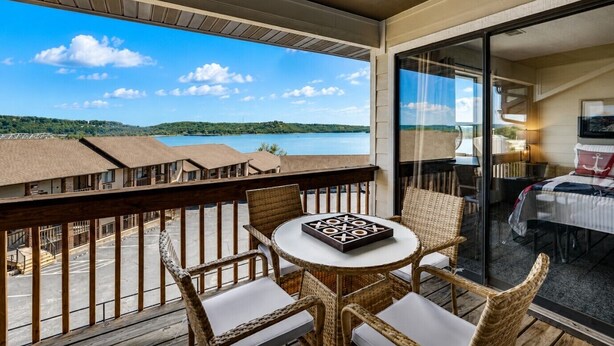 A balcony with wicker chairs, a small table, and a view of a lake and buildings. - Captain`s Quarters at Kimberling Crossing (Kimberling City)