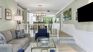 Un salon spacieux avec une table basse en verre, un fauteuil bleu et un canapé gris. Au fond, on aperçoit un coin repas avec une rambarde blanche.