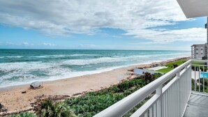 Blick auf einen Strand vom Balkon aus, mit Pool und einem Gebäude im Hintergrund.