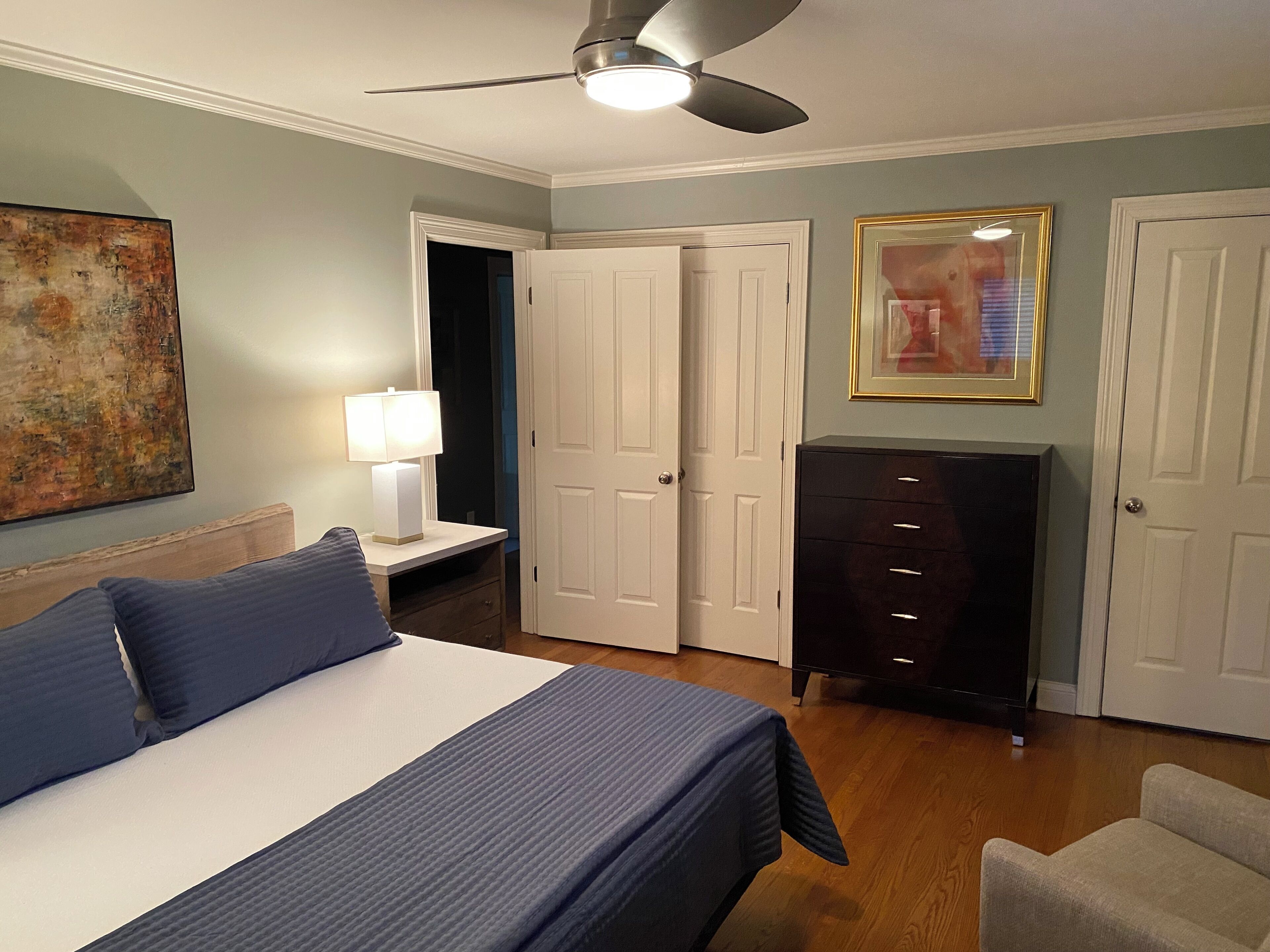 A bedroom with a bed, a dresser, a desk with a lamp, a ceiling fan, and a painting on the wall.