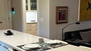 A modern kitchen with a white countertop, two bar stools, a dining table set with plates and a mug, and a painting on the wall. - Murfreesboro Mid-Century Modern with AC, WiFi (Murfreesboro)