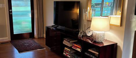 A living room with a flat-screen TV, wooden entertainment unit, bookshelves, a rug, and a window with blinds.