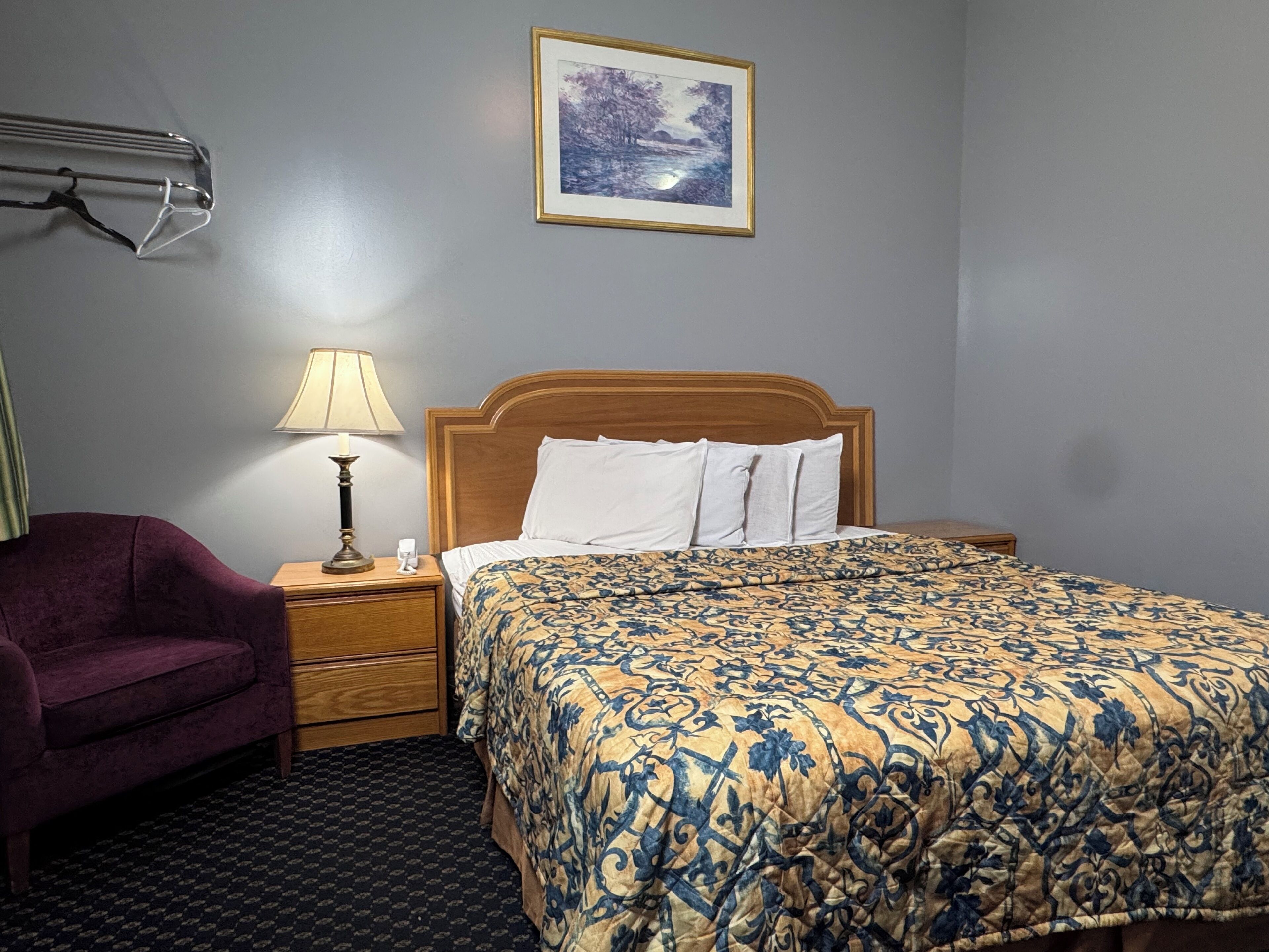Una habitación de hotel con una cama de madera, un edredón estampado, una mesita de noche con una lámpara, una silla morada y un cuadro en la pared.
