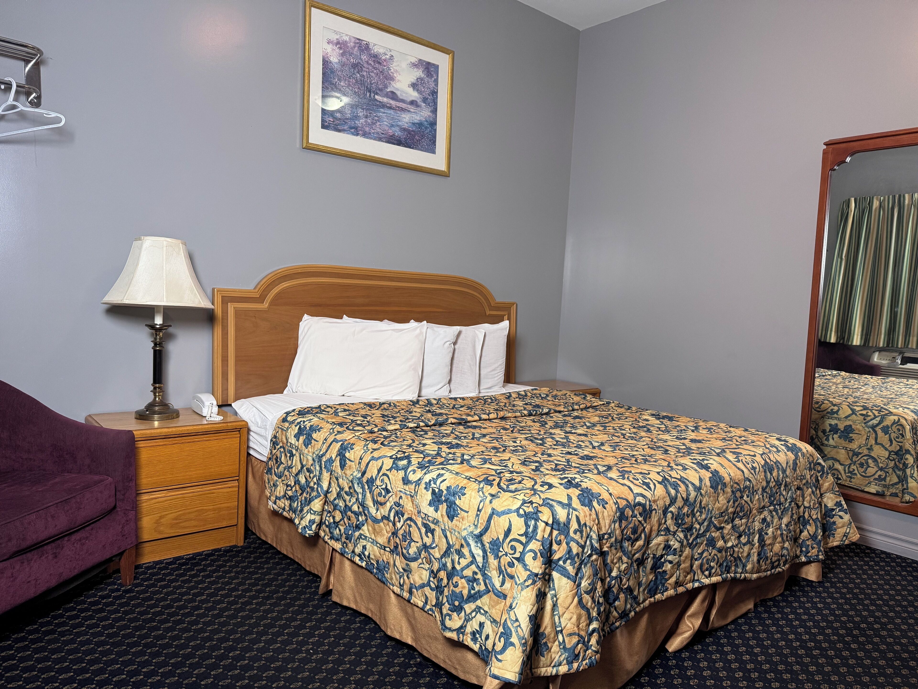 A hotel room with a bed, a nightstand with a lamp, a mirror, a purple chair, and a framed picture on the wall.