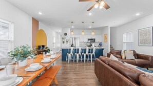 Un salon moderne avec un canapé en cuir marron, un coin repas avec une table en bois dressée pour quatre personnes, et une cuisine en arrière-plan.