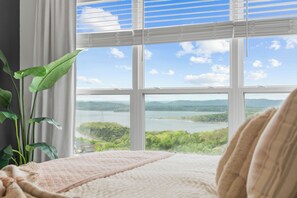 A bedroom with a large window offering a view of a lake and hills.