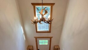 Un escalier en bois surmonté d’un lustre, une porte avec une fenêtre et un luminaire fixé au mur.