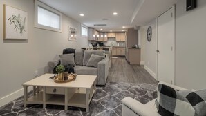 A modern living room with a gray sofa, a coffee table, and a rug. There is a kitchen in the background with wooden cabinets and a refrigerator. - Fully Reno'd 2 Bedroom, 2 Bath Legal Basement Suite. Heart of the Southside (Lethbridge)