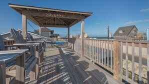 A wooden deck with a covered area, a sign reading "JACKDOG EST 1979", and a view of the beach and houses in the background. - 7041- Duck Dog - Rodanthe Beach Cottage | Ocean Views (Rodanthe)