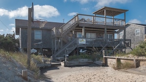 A beachfront property with a multi-level deck, a staircase leading to the upper floor, and a sign indicating a resort realty business. - 7041- Duck Dog - Rodanthe Beach Cottage | Ocean Views (Rodanthe)
