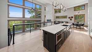 A spacious kitchen with a large island, high ceilings, and large windows offering views of the outdoors. - Retreat at Clear Creek ~ Outdoor Pool & Pickleball/Basketball Court! (Ellijay)