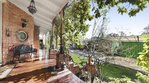 A covered patio with a brick wall, a bench, and a view of a garden and green fence.