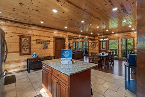 A wooden cabin interior with a kitchen, dining area, and a large window overlooking greenery.
