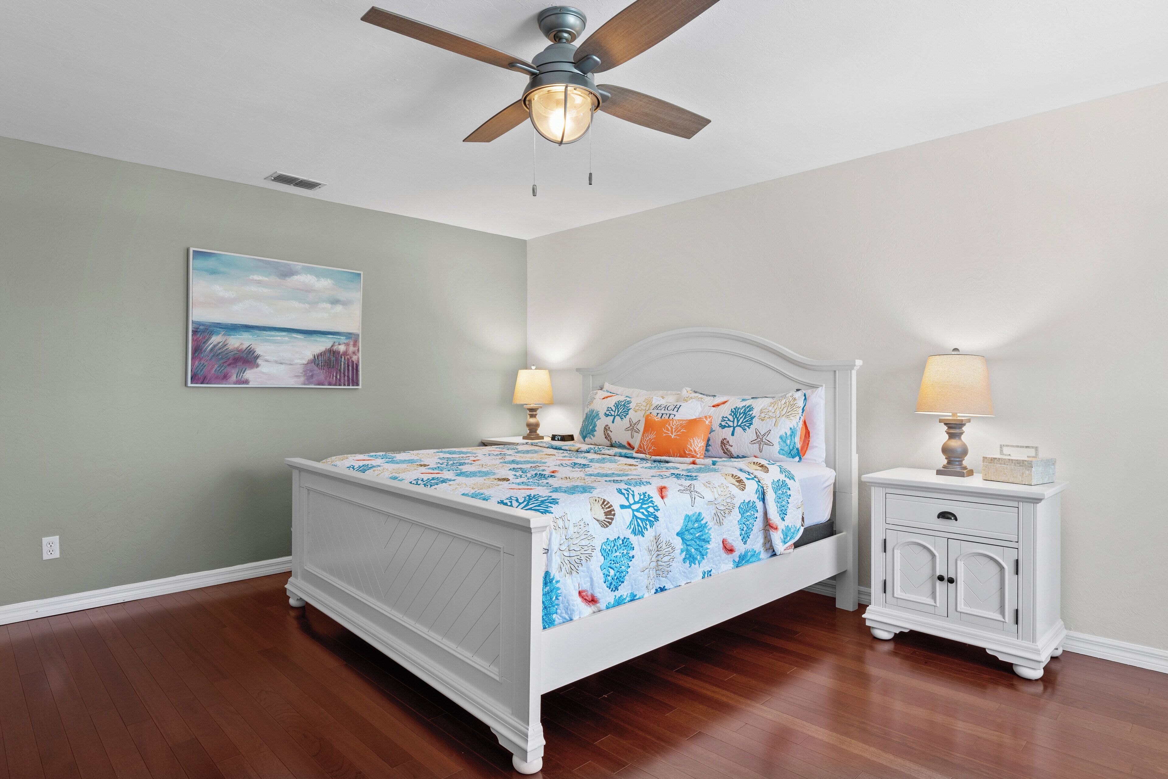 Un dormitorio con cama, mesitas de noche, ventilador de techo y un cuadro en la pared.