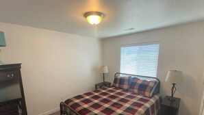 A bedroom with a bed, two bedside tables, a window with blinds, a ceiling light fixture, and a wall-mounted shelf.
