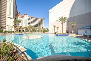 A hotel pool area with clear blue water, palm trees, and a large building in the background.