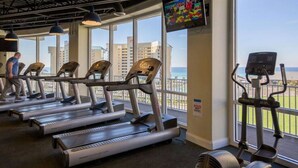 A gym with treadmills and an elliptical machine, large windows with a view of the ocean and buildings, and a television mounted on the wall. - Palm Tree Paradise – Free Beach Chairs & Umbrella, 5 Pools, Beach Access | Laketown Wharf 1531 (Panama City Beach)