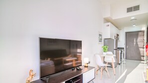 Ein modernes Wohnzimmer mit großem Flachbildfernseher, einem Essbereich mit kleinem Tisch und Stühlen sowie einer Küche im Hintergrund.