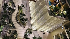 Un vestíbulo moderno de hotel con una escalera espiral central, una zona de piscina con iluminación azul y abundante vegetación circundante.