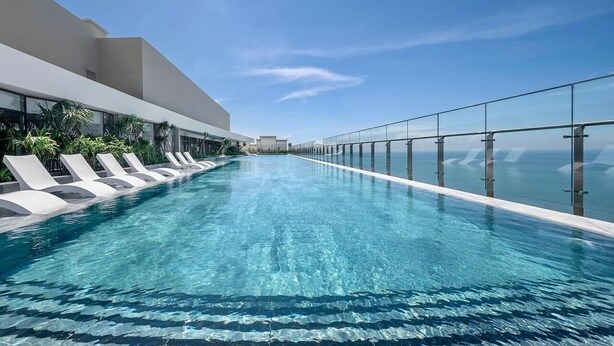 Una piscina infinita moderna con agua azul cristalina, rodeada de sillones blancos y barandillas de vidrio, con vistas a un paisaje urbano.