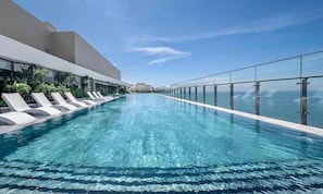 A modern infinity pool with clear blue water, surrounded by white lounge chairs and glass railings, overlooking a cityscape. - Bin's House in The Song Vung Tau (Ho Chi Minh City)