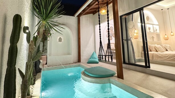 A swimming pool with a small waterfall feature, surrounded by cacti and a potted plant. There is a cozy seating area with cushions and a hanging light fixture. - Stunning 1 bedroom villa in Seseh area (Bali)