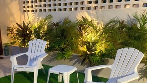 Eine Terrasse mit zwei weißen Adirondack-Stühlen und einem kleinen Tisch, umgeben von Kunstgrashalm und verschiedenen Pflanzen.