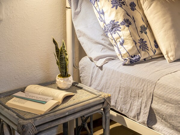 En säng med randigt sänglinne och blommiga kuddar, en liten krukväxt och en öppen bok på ett metallbord.
