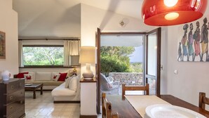 A living room with a sofa, a dining table with a plate, and a lamp. There is a window with curtains and a view of greenery outside. - Vacation home Giulia by Interhome (Portobello)