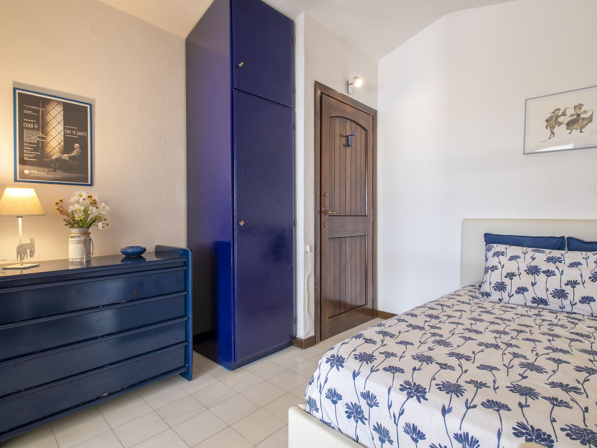 A bedroom with a bed, a dresser, a wardrobe, a door, and a wall-mounted lamp.
