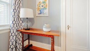Una consola de madera con un jarrón blanco y una lámpara, un cuadro de flores en la pared y una cortina estampada.