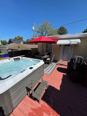 A hot tub on a wooden deck with a red umbrella, a barbecue grill, and a covered patio area.