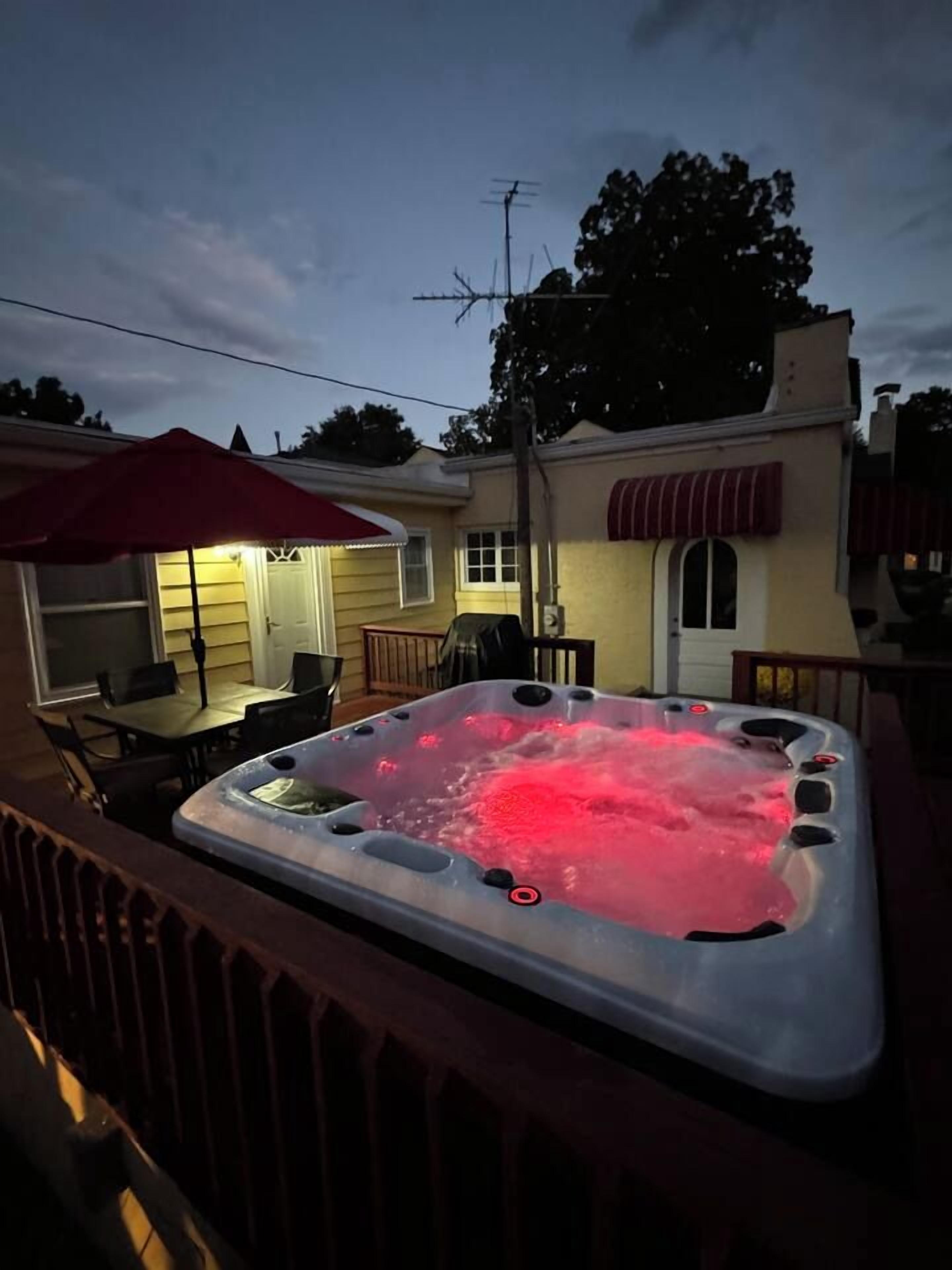 A hot tub on a deck with a patio table and chairs, a covered outdoor seating area, and a house in the background.