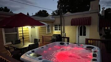A hot tub on a deck with a patio table and chairs, a covered outdoor seating area, and a house in the background.