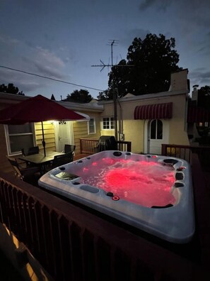 A hot tub on a deck with a patio table and chairs, a covered outdoor seating area, and a house in the background.