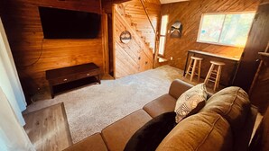 L’intérieur chaleureux d’un chalet, avec un revêtement mural en bois, un canapé, un banc, un téléviseur fixé au mur et un petit coin bar.